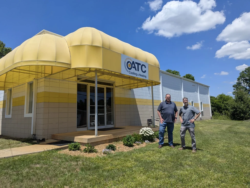Two men standing on a grassy lawn in front of the ATC Cooling & Heating building, which features a large, curved yellow awning over the main entrance.